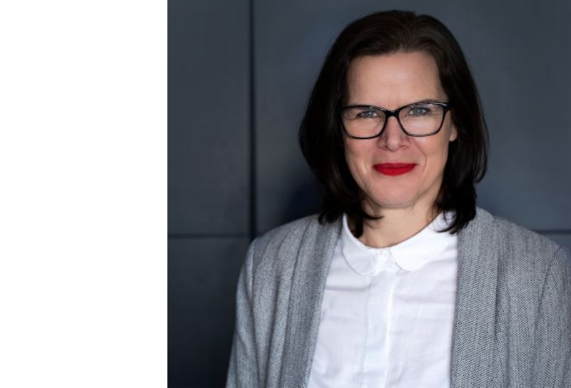 Person mit schulterlangen schwarzen Haaren, mit einer Brille. Sie trägt ein weißes Hemd mit einem grauen Blazer. 