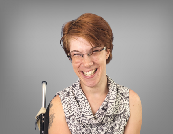 Natascha Toman smiles at the camera. She has red short hair, wears glasses and uses a wheelchair.