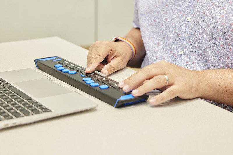 zwei Hände bedienen eine Braille Tastatur, im Anschnitt ist ein Computer zu sehen