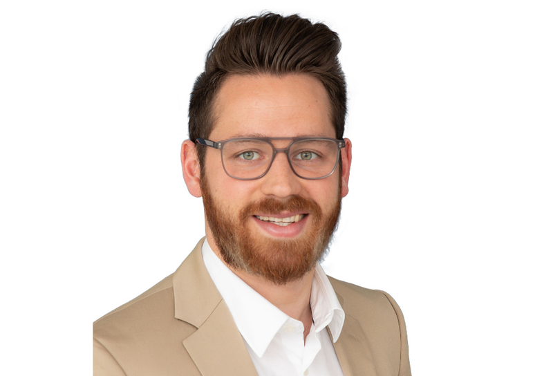 Die Person hat kurze braune Haare, eine Brille und einen Bart. Er trägt ein beiges Jackett und ein weißes Hemd. 
