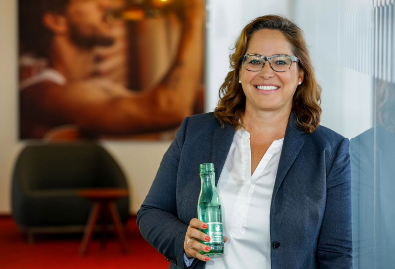Die Person hat schulterlange braune Haare und eine Brille. Sie trägt einen grauen Blazer mit einem weißen T-Shirt. Sie hält eine Glasflasche in der Hand.
