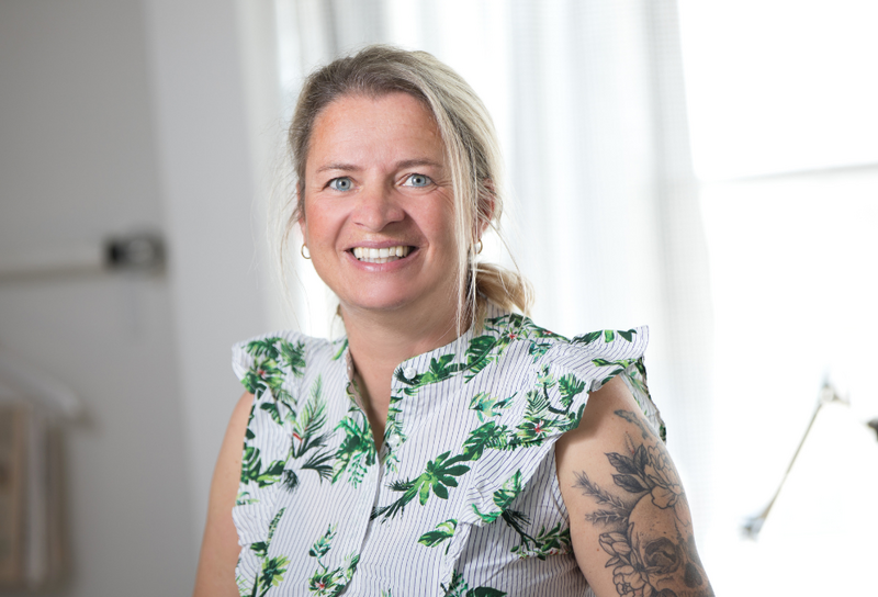 Die Person hat blonde lange Haare zu einem Zopf zusammen gebunden. Sie hat ein Tattoo am Oberarm und trägt eine weiße Bluse mit Muster.