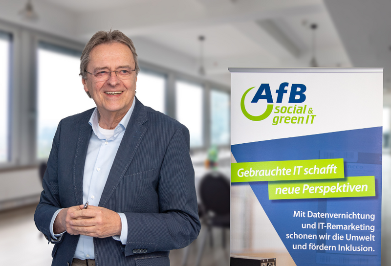 Person mit kurzen grauen Haaren und einer Brille. Er trägt ein helles Hemd mit einem grauem Blazer. Im Hintergrund ist ein ein AfB Banner. 