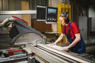 Eine Frau mit mittellangen, zusammengebundenen Haaren in blauer Handwerkshose arbeitet an einer Kreissäge. Sie trägt einen professionellen Gehörschutz und blickt konzentriert auf das Holz, das sie gerade verarbeitet.