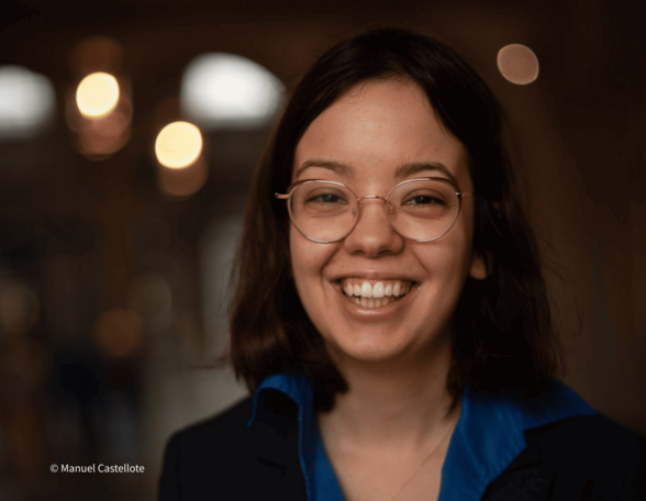 Foto einer Person mit schulterlangen, braunen Haaren und Brille. Sie lacht herzlich in die Kamera, trägt einen dunklen Blazer, ein blaues Hemd und eine Goldkette. 