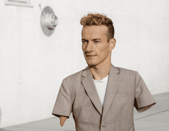 Eine Person mit kurzen lockigen Haaren steht vor einer hellen Betonwand und trägt ein beigefarbenes Sakko über einem weißen Shirt. Die Person hat keine Arme und Beine.