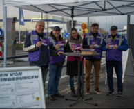 Fünf Personen unter einem Pavillon, tragen violette Westen und halten violette Flyer, Stehtisch mit Flyer Stapel.