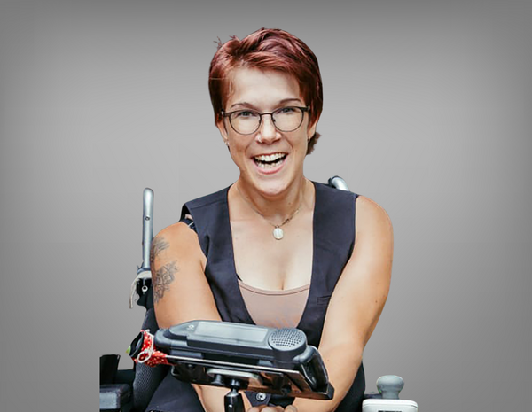 Natascha Toman lächelt in die Kamera. Sie hat rote kurze Haare, trägt eine Brille und nutzt einen Rollstuhl.