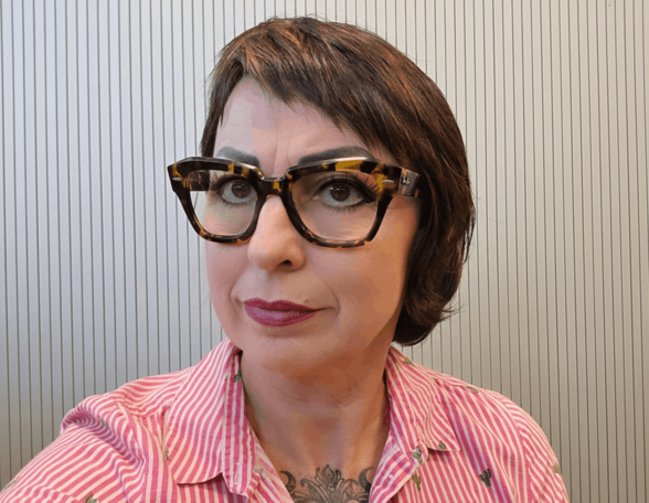 Person mit braunen, kinnlangen Haaren und auffälliger Brille mit Muster steht vor einer Bretterwand. Sie trägt eine karierte Bluse und roten Lippenstift.