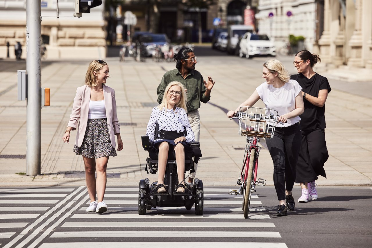 4 women and a man cross a zebra crossing. One person pushes a bicycle, one person uses a wheelchair.