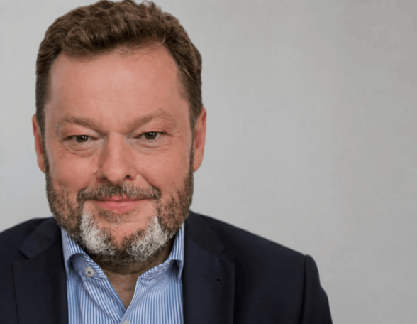 Person mit kurzem, brünettem Haar und graumeliertem, kurzem Bart. Sie trägt ein blaues Hemd, der oberste Knop offen und ein dunkles Sakko.