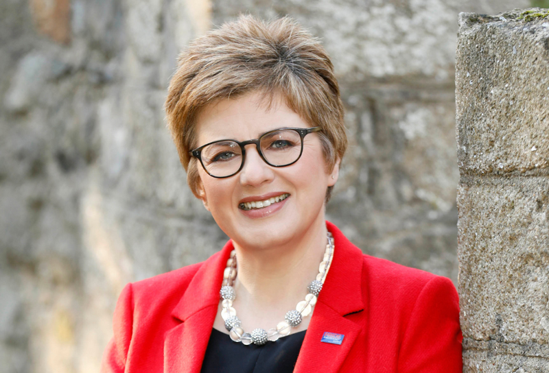 Die Person hat kurze braune Haare und trägt Schmucke, wie auch eine Brille. Sie trägt eine schwarzes T-Shirt und einen roten Blazer.