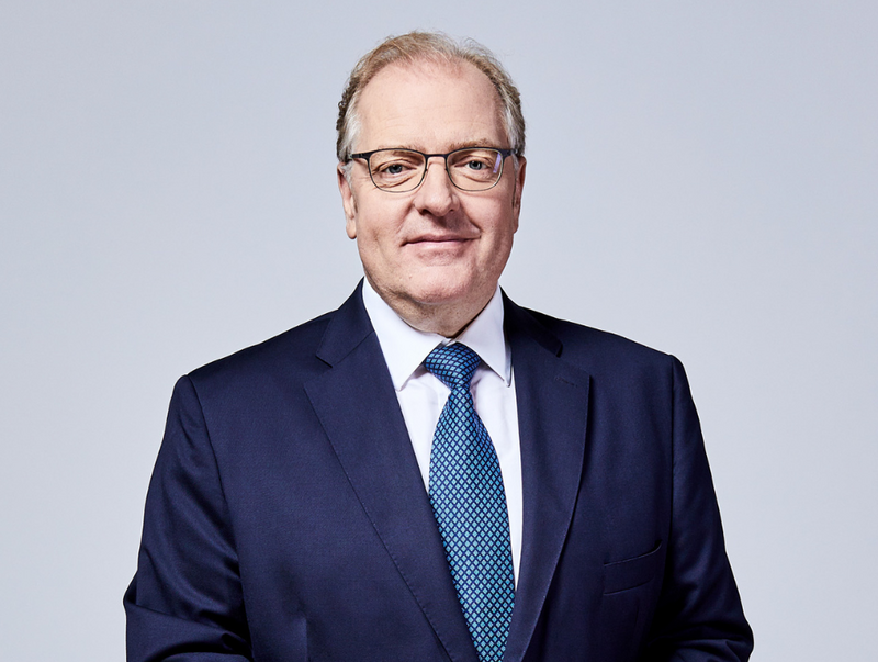 Porträt von Helmut Bernkopf. Er hat kurze weiße Haare. Er trägt eine Brille, ein dunkelblaues Sakko mit einem weißen Hemd und einer blau gemusterten Krawatte.