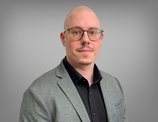 Portrait von Lukas Sukal. Er hat eine Glatze, einen Bart, eine Brille und ein Nasenpiercing. Er trägt ein schwarzes Hemd mit grauem Sakko.