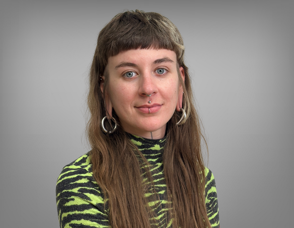 Portrait von Lena Cebul. Sie hat lange braune Haare und Piercings. Sie trägt ein schwarz grün gemustertes T-Shirt.