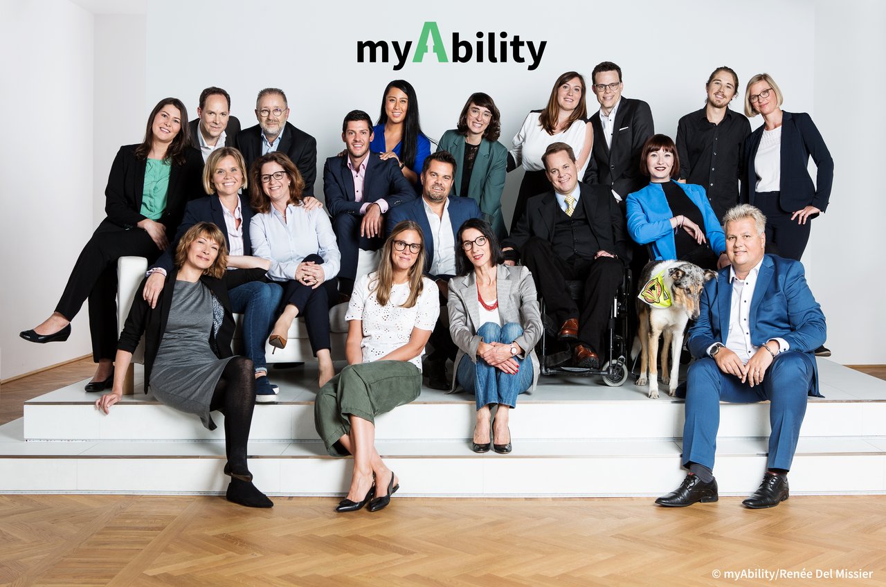 Das Team von myAbility in die Kamera schauend, etwa 20 Personen mit und ohne Behinderungen in Business-Kleidung