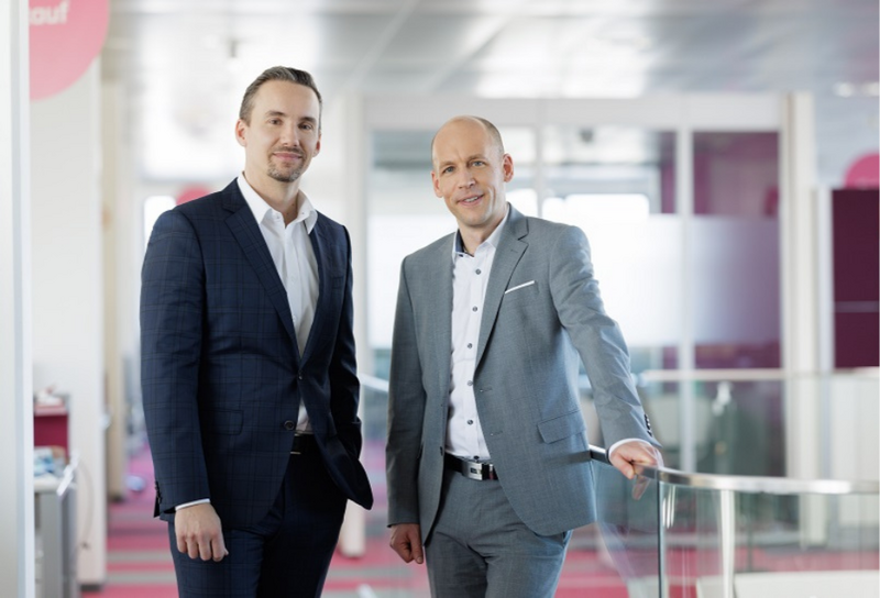 Links sthet ein Herr mit weißen Hemd und schwarzem Sakko. Rechts steht ein Herr mit weißem Hemd und grauem Sakko