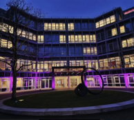 Mehrstöckiges Bürogebäude am Abend, Erdgeschoss in Violett beleuchtet, Innenhof mit Skulptur aus zwei schwarzen Ringen
