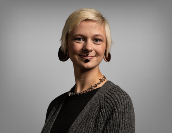 Portrait von Annika Candy. Sie hat kurzes blondes Haar und und trägt Piercings. Sie trägt ein schwarzes Shirt mit einer grauen Weste.