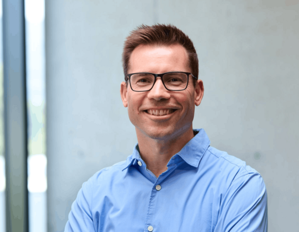 Peron mit kurzem, braunem Haar und Brille steht lächelnd vor einer Hauswand. Sie trägt ein hellblaues Hemd.
