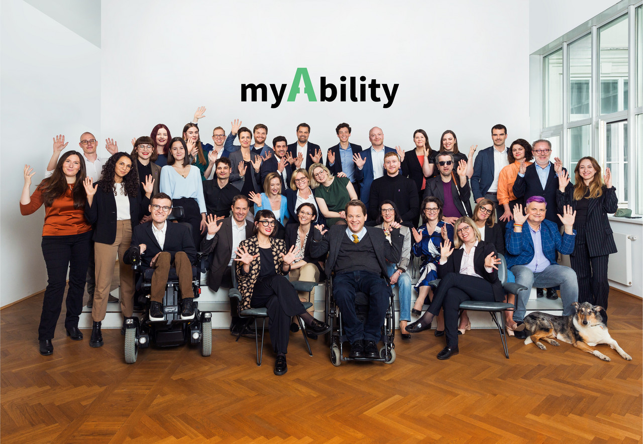 Teamfoto myAbility März 2023. 37 Personen mit und ohne Behinderungen applaudieren in Gebärdensprache und lächeln. myAbility Logo auf weißer Wand im Hintergrund