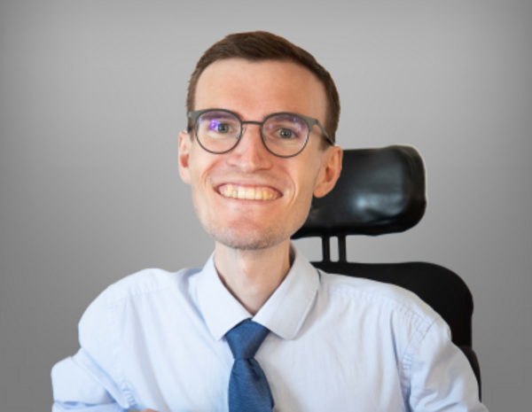 Portrait of Lukas Leitner, male person with short brown hair in blue shirt and blue tie in the background neckrest of an electronic wheelchair