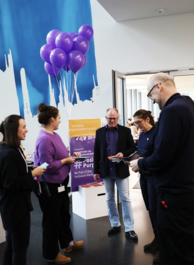 5 Personen im Gespräch. In der Mitte ein Stand mit #PositivelyPurple Plakat und lila Luftballons.