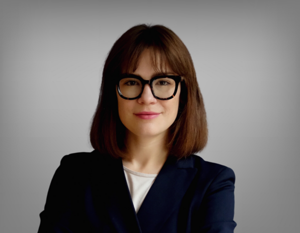 Portrait von Anna Herzog, sie hat schulterlange braune Haare und trägt eine Brille. Sie hat ein weißes T-Shirt mit einem schwarzen Blazer an.