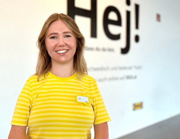 Person mit schulterlangen blonden Haaren und gelbem T-Shirt steht vor einer Wand in einem IKEA Möbelhaus.