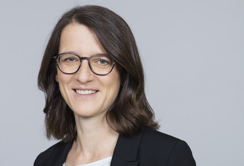 Die Person hat schulterlange braune Haare und eine Brille. Sie trägt ein weißes T-Shirt mit einem schwarzen Blazer.