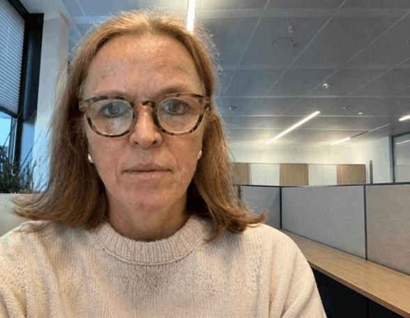 Person mit schulterlangen, blonden Haaren und Brille in einem Büro. Sie trägt einen grauen, hochgeschlossenen Pullover.