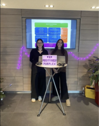Zwei Personen hinter einem Stehtisch mit zwei Laptops, violette Lichterkette im Hintergrund. Schild: „♥ # ♥ POSITIVELY PURPLE ★“. Bildschirm: „Agenda Gesundheitstage“.
