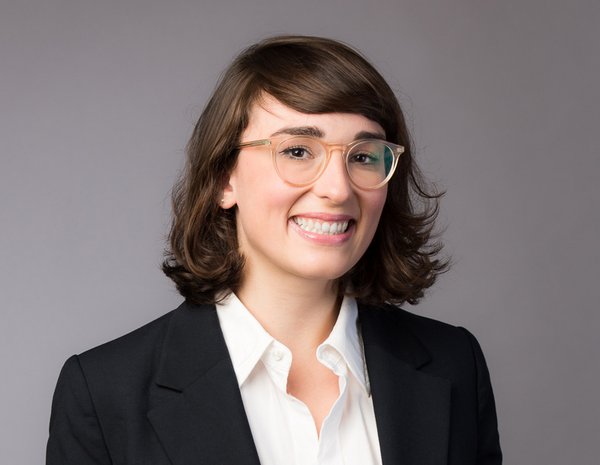 Portrait of Fides Raffel, female person with brown shoulder-length hair and glasses. She wears a white blouse and a black blazer.