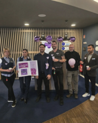 6 Personen mi lila Luftballons. Eine Person hält ein Plakat mit #PositivelyPurple Grafiken und Informationen