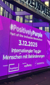 Großer violetter Screen an einem Gebäude mit den Texten „#PositivelyPurple“, „Part of the Inclusion Revolution!“, „3.12.2025“, „Internationaler Tag der Menschen mit Behinderungen“, „Powered by PurpleSpace“, „Supported by myAbility“ und „Rheinmetall“.