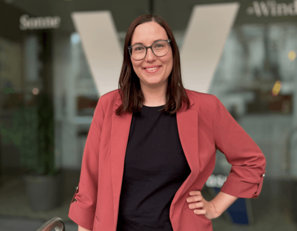 Person vor eine Glastüre mit dem Buchstaben V darauf. Sie hat schulterlange braune Haare, trägt Brille, ein rotes Sakko und eine schwarze Bluse.