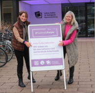 Zwei Personen stehen mit einem Plakat vor dem Gebäude der Leibniz- Institut für ökologische Raumentwicklung. Auf dem Plakat steht: "#PositivelyPurple. Aktiv Farbe bekennen. Wir stehen ein für eine inklusive und barrierefreie Arbeitswelt."