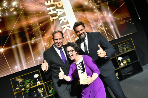 Michael Aumann, Stefanie Kirwald und Wolfgang Kowatsch nach der Preisverleihung. Sie strecken die Daumen in die Luft, in Stefanies Händen die Trophäe mit Aufschrift „The Spark“. Ihre Gesichter strahlen fröhlich. Stefanie trägt ein violettes Kleid, das farblich an die #PurpleLightUp Kampagne erinnert.