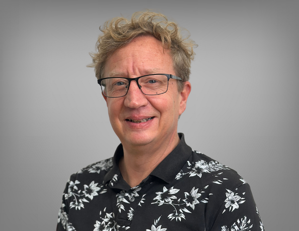 Portrait von Oswald Schwarz. Oswald hat kurze lockige helle Haare und trägt eine Brille. Er trägt ein schwarzes T-Shirt mit weißen Blumen.