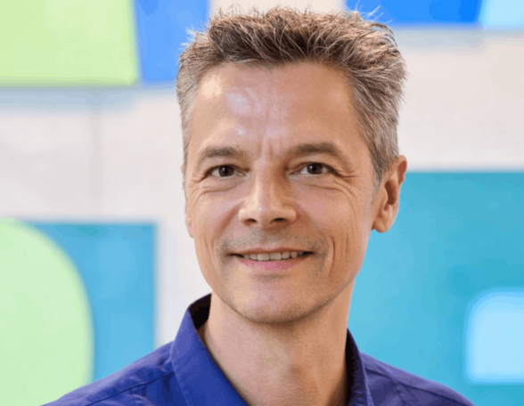 Person mit kurzen, graumelierten Haaren und dunkelblauem Hemd vor einem grün-blauen Hintergrund.