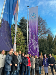 Gruppe von Menschen steht im Freien vor Fahnenmasten; daneben hängt ein violettes Banner mit einem Rollstuhl-Piktogramm und einem Emblem zum Internationalen Tag der Menschen mit Behinderungen.