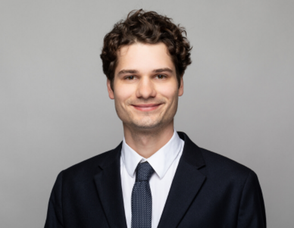 Portrait von Laurenz Miller, männlich gelesene Person mit kurzem schwarzem Haar und schwarzem Anzug und blauer Krawatte