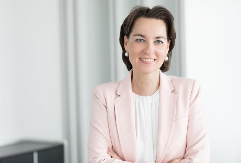 Die Person hat schulterlange braune Haare und trägt Ohrringe. Sie trägt eine weiße Bluse mit einem rosafarbenen Blazer.