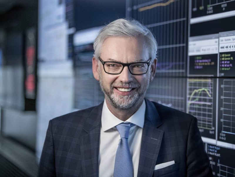 Porträt von Michael Strugl mit weißen kurzen Haaren und Bart sowie Brille. Er trägt einen dunklen Anzug, ein weißes Hemd und eine hellblaue Krawatte. Im Hintergrund Bildschirmen mit Grafiken und Zahlen.