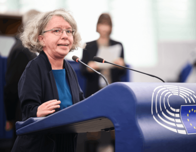 Eine Person mit lockigen, grauen Haaren und Brille steht an einem Podium mit Mikrofonen. Man sieht das Logo des Europäischen Parlaments.