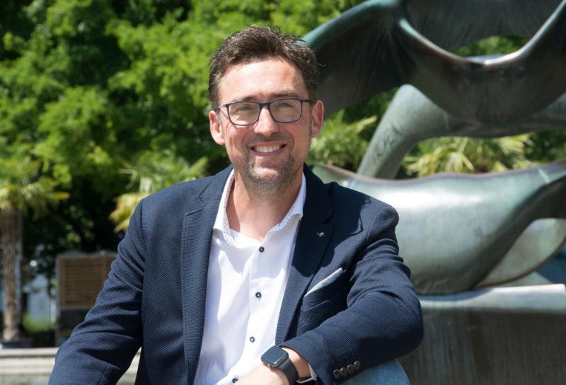 Person mit kurzem, braunen Haar, trägt Brille, ein blaues Sakko, ein weißes Hemd und eine Armbanduhr. Die Person lächelt in die Kamera. Im Hintergrund sieht man Bäume und einen Teil einer Statue.