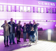 Eine Gruppe von Menschen vor dem lila beleuchteten Siemens Gebäude formen ihre Hände zu einem Herz.
