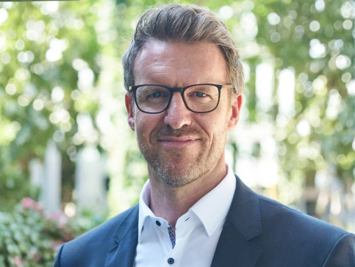 Porträt eines lächelnden Mannes mit Brille, dunklem Sakko und weißem Hemd vor unscharfem grünen Hintergrund im Freien.