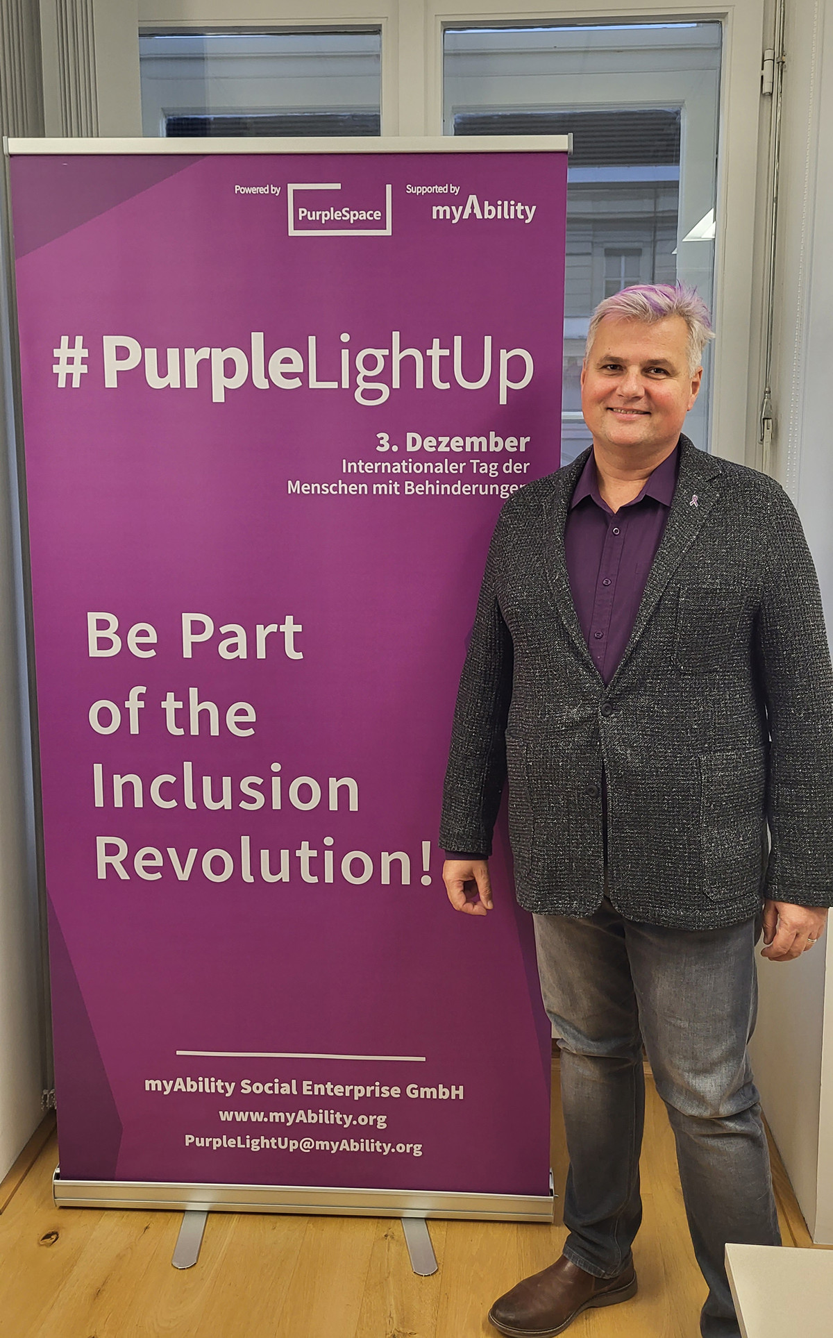 Robert Öllinger mit lila gefärbtem Haar, neben einem lila Roll-Up "#PurpleLightUp - 3. Dezember, Internationaler Tag der Menschen mit Behinderungen"
