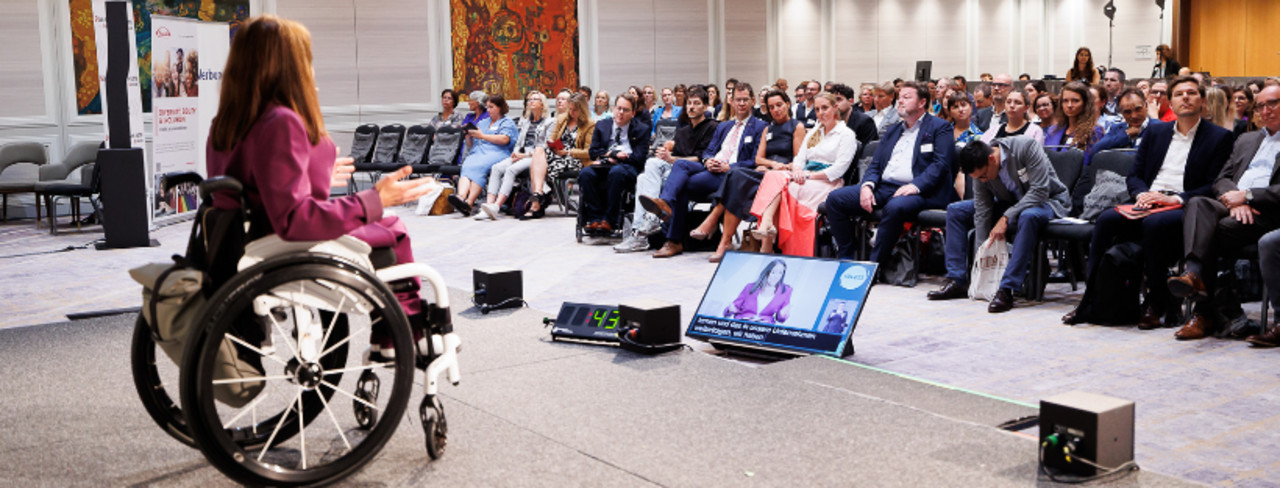Moderatorin im Rollstuhl auf der Bühne, Publikum im Hintergrund